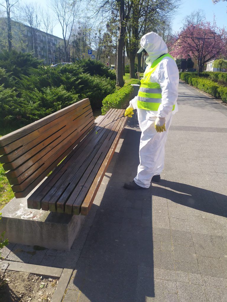 Pracownik komunalny ubrany w specjalny kombinezon dezynfekuje ławkę