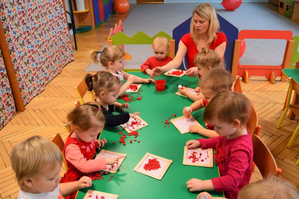 Maluchy siedzą przy stole i wraz opiekunką rysują malują czerwone serduszka