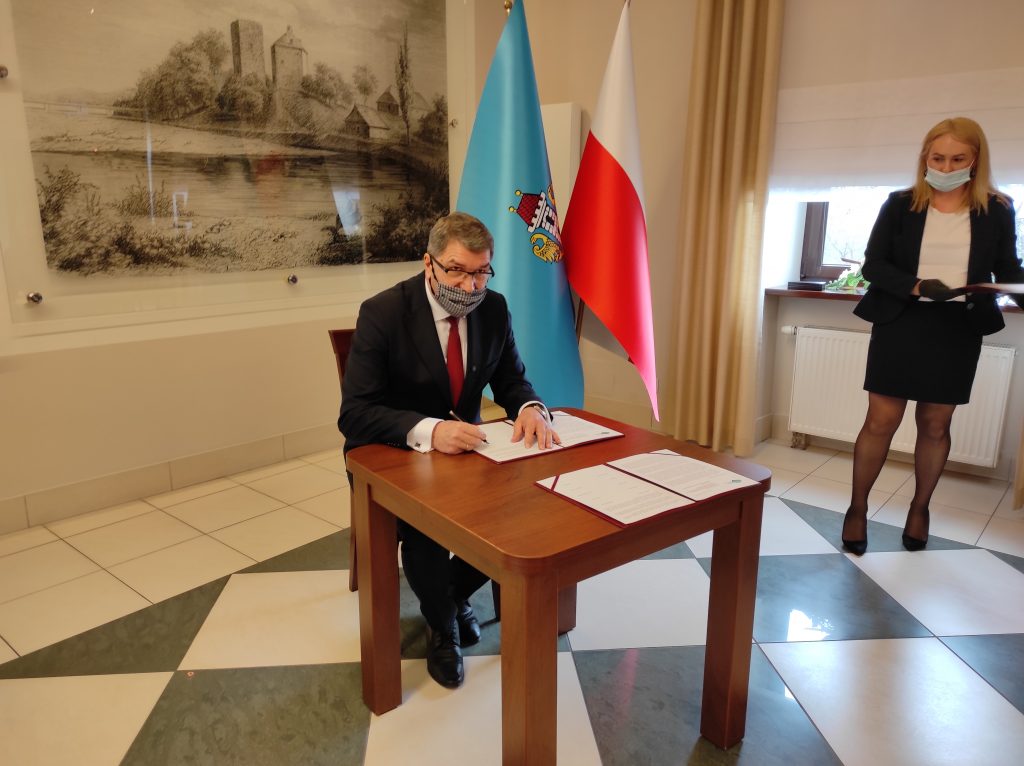 Prezydent Oświęcimia siedzi przy stole i podpisuje dokument. W tle  grafika wzgórza zamkowego i zamku od strony Soły, obok pióropusz z flagami: Polski, Oświęcimia i Unii Europejskiej