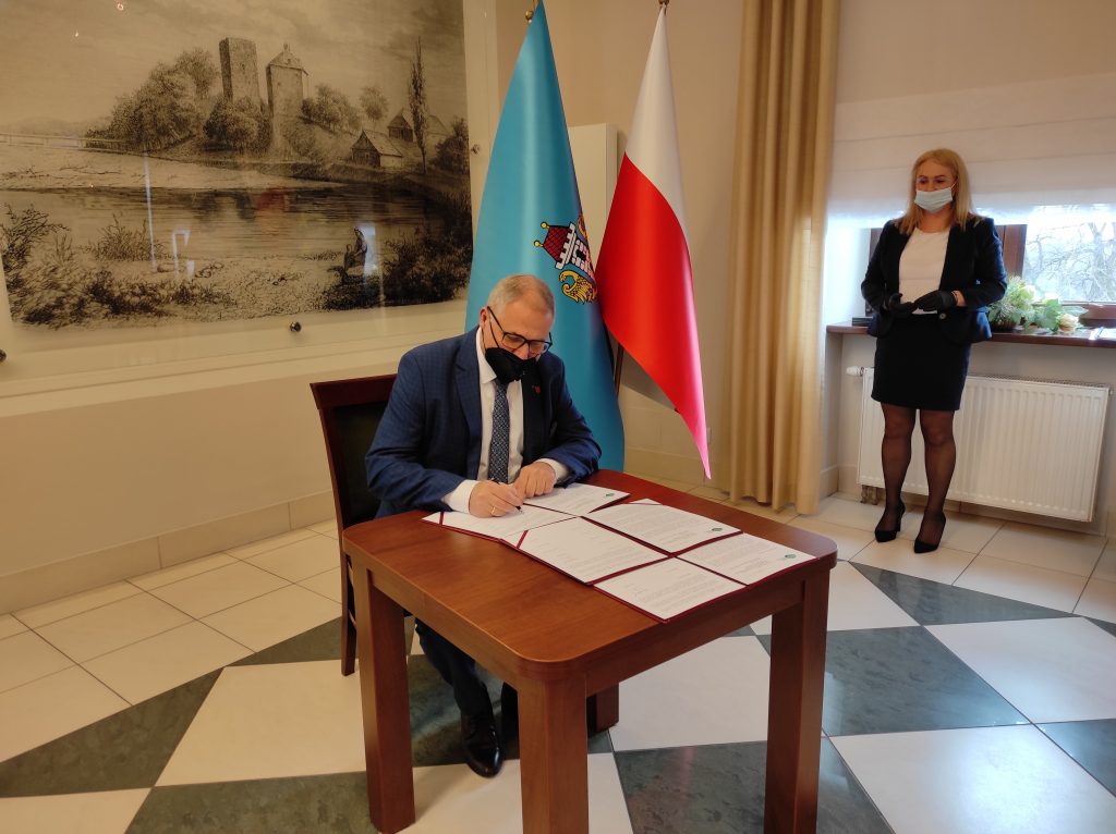 Burmistrz Andrychowa siedzi przy stole i podpisuje dokument. W tle  grafika wzgórza zamkowego i zamku od strony Soły, obok pióropusz z flagami: Polski, Oświęcimia i Unii Europejskiej