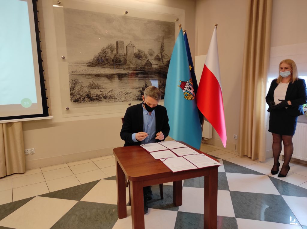 Burmistrz Brzeszcz siedzi przy stole i podpisuje dokument. W tle  grafika wzgórza zamkowego i zamku od strony Soły, obok pióropusz z flagami: Polski, Oświęcimia i Unii Europejskiej