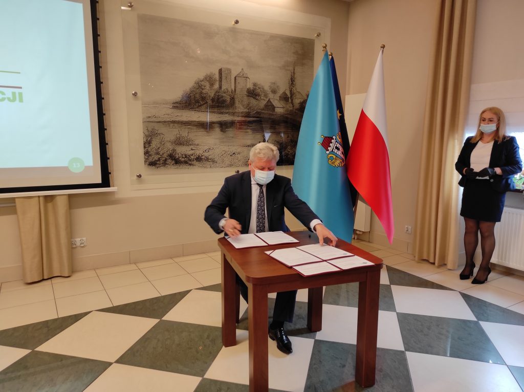 Burmistrz Chełmka siedzi przy stole i podpisuje dokument. W tle  grafika wzgórza zamkowego i zamku od strony Soły, obok pióropusz z flagami: Polski, Oświęcimia i Unii Europejskiej