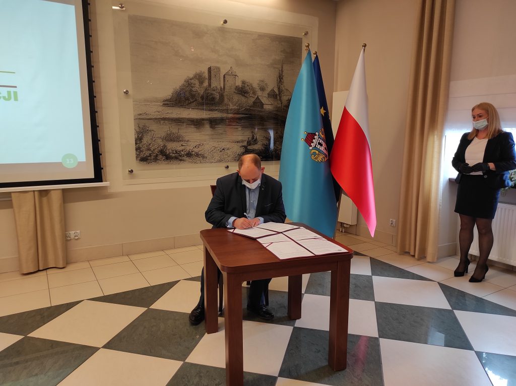 Burmistrz Kęt siedzi przy stole i podpisuje dokument. W tle  grafika wzgórza zamkowego i zamku od strony Soły, obok pióropusz z flagami: Polski, Oświęcimia i Unii Europejskiej
