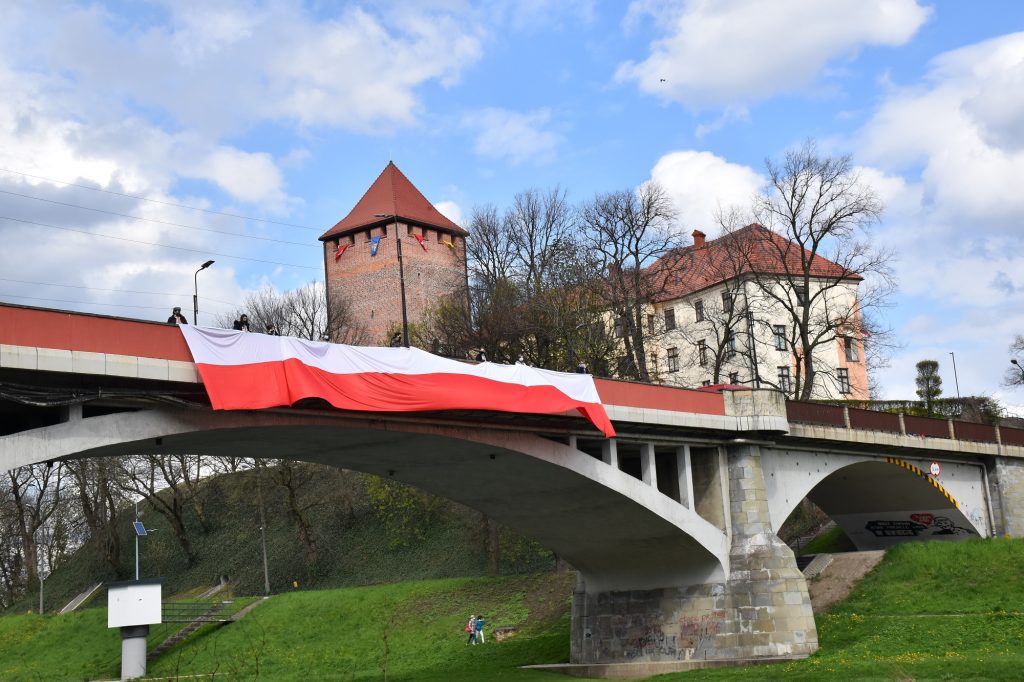 Flaga Polski na moście nad rzeką Sołą trzymana przez harcerzy. Niebo pochmurne a wokoło dużo zieleni. W tle zamek i wieża. 