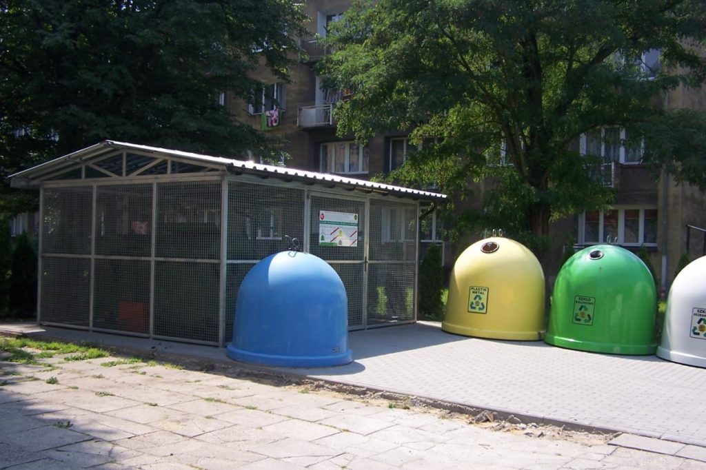 Dzwony do segregacji odpadów w kolorze niebieski, żółtym i zielonym stoją przy wiacie śmietnikowej