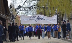 Grupa młodych ludzi z banerem Marsz Żywych  2022 przechodzi przez bramę z napisem Arbeit Mach Frei