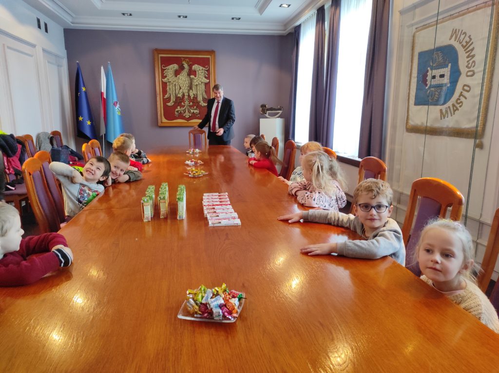 Dzieci siedzą przy stole, prezydent stoi. Na stole słodki poczęstunek i napoje