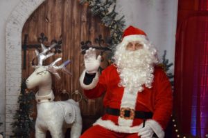 Św. Mikołaj obok ceramiczny renifer. Mikołaj ma uniesioną rękę w geście pozdrowienia.
