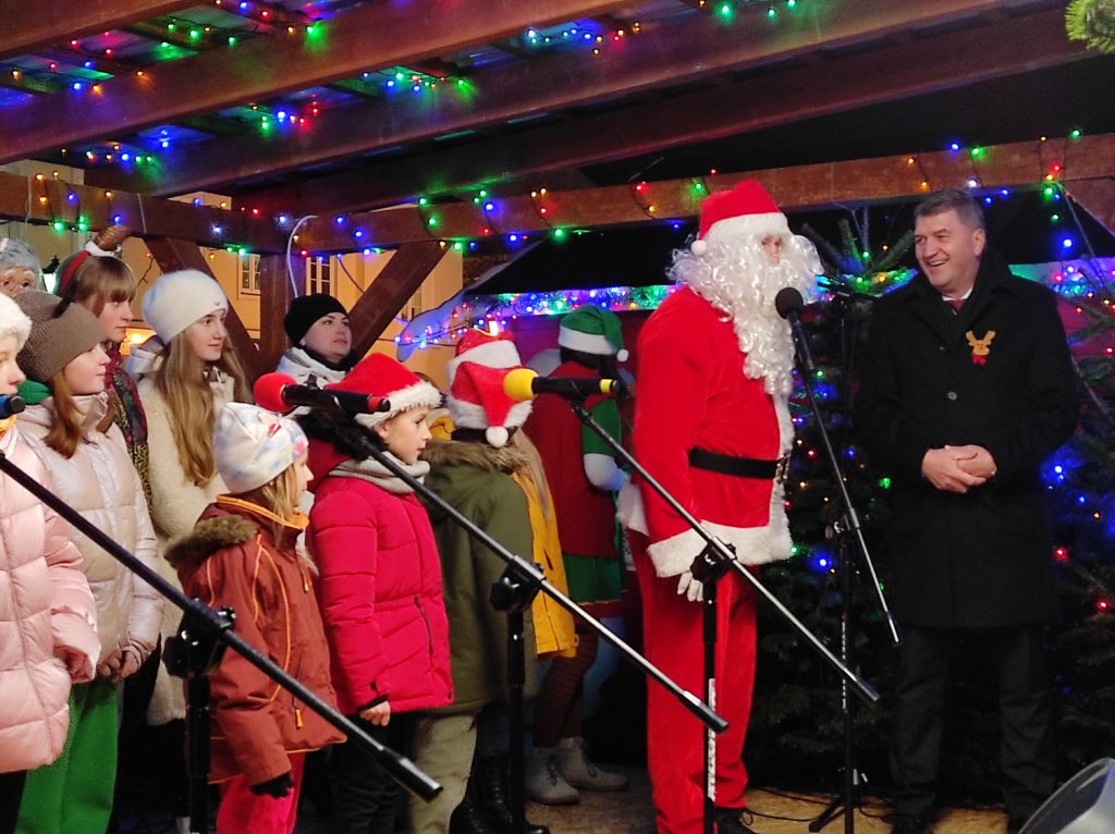 Święty Mikołaj pozdrawia ze sceny dzieci. Obok stoi prezydent Oświęcimia oraz chór dzieci