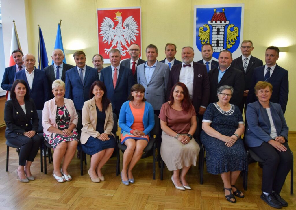Na fotografii 21 radnych, w pierwszym rzędzie siedzą radne, za nimi stoją radni. W tle herb Polski, miasta i flagi