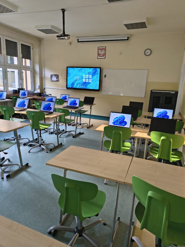 Pracownia sztucznej inteligencji - zielone krzesła, biurka na których znajdują się srebrne laptopy, na ścnianie znajduje się monitor interaktywny